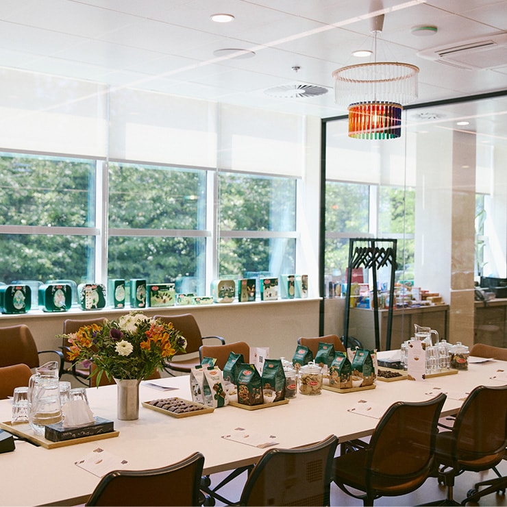 Salle de réunion lumineuse avec table décorée de fleurs, grandes fenêtres et chaises marron avec de Delacre biscuits sur la table.